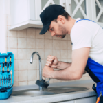 Plumber using a plunger to unclog a kitchen drain with a toolbox open beside him.