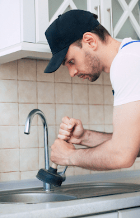 Plumber using a plunger to unclog a kitchen drain with a toolbox open beside him.