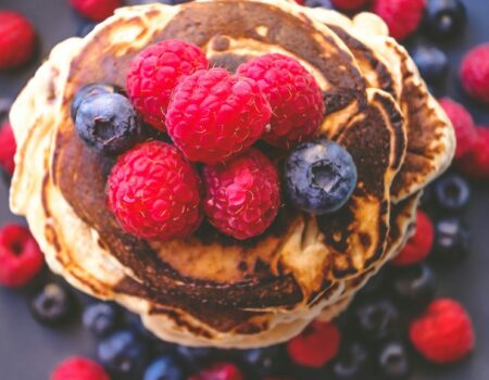 baking without eggs: A stack of fluffy vegan pancakes. topped with fresh raspberries and blueberries, surrounded by additional berries on a dark plate.
