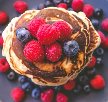 baking without eggs: A stack of fluffy vegan pancakes. topped with fresh raspberries and blueberries, surrounded by additional berries on a dark plate.