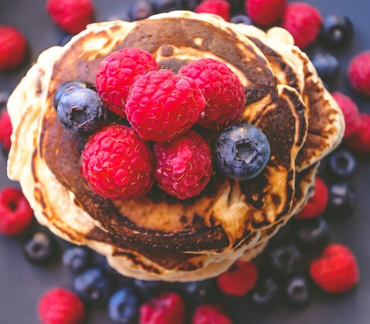 baking without eggs: A stack of fluffy vegan pancakes. topped with fresh raspberries and blueberries, surrounded by additional berries on a dark plate.